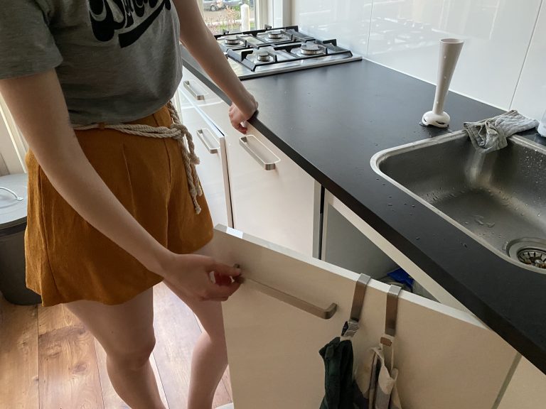 Person standing in front of the kitchen counter, opening the cupboard under the sink.